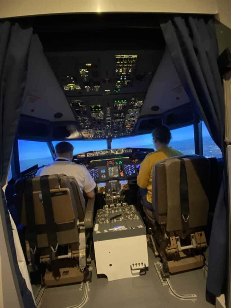 Susan is sat in the pilot chair of a flight simulator ready to the land the plane with the help of a pilot as part of the aviation career day 